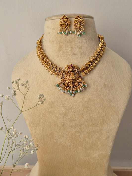 Handcrafted traditional gold-plated temple jewelry necklace featuring an intricate Goddess Lakshmi motif pendant with green beads and pearl cluster fringes, paired with matching stud earrings By naitri.in