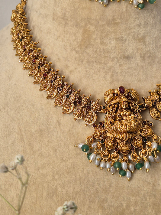 Antique gold-polished brass necklace set showcasing structured goddess with floral metalwork, ruby-red and emerald-green stone accents, and hanging seed pearls from the Naitri Golden Ethnic collection by naitri.in