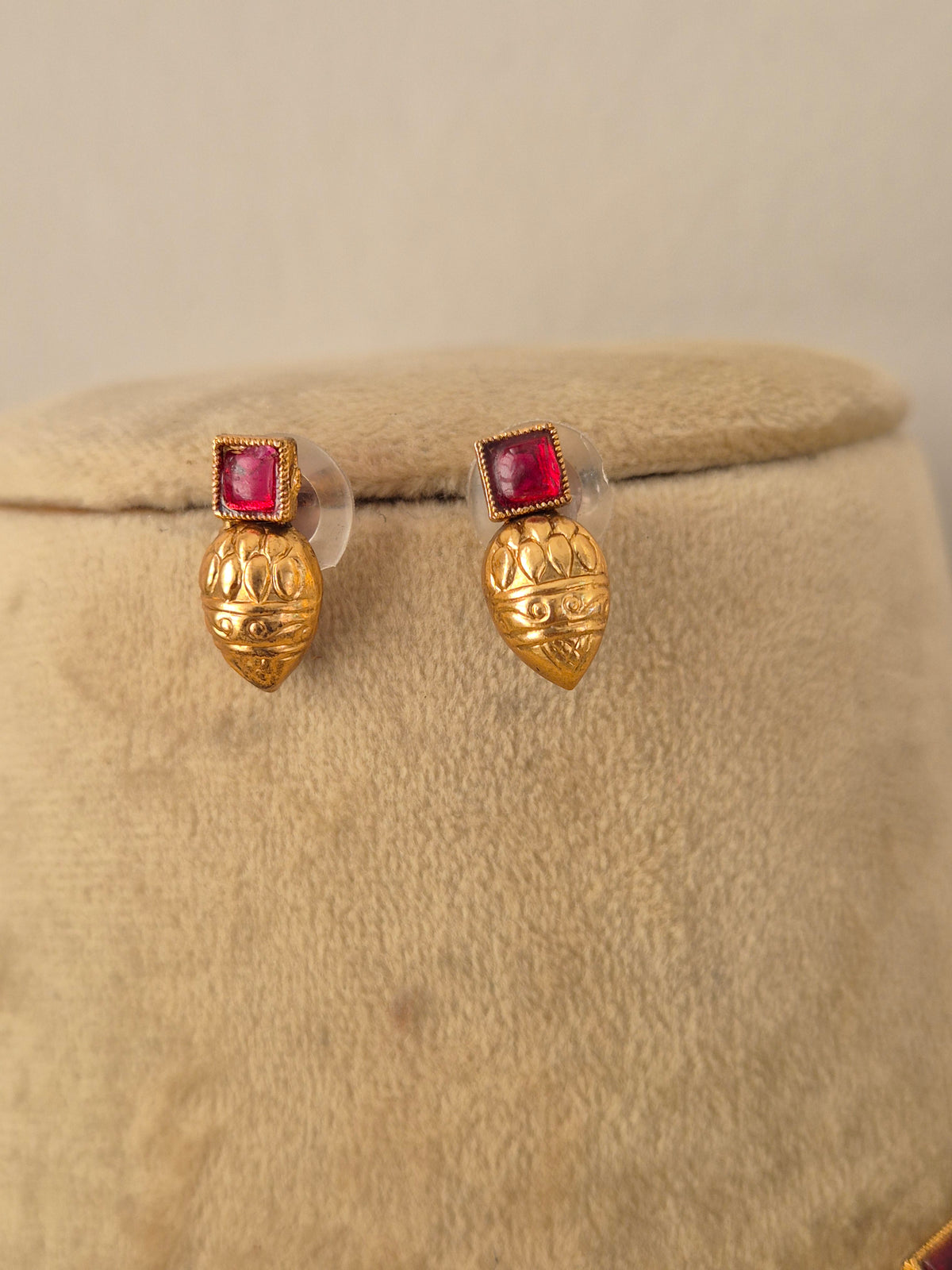 A pair of gold post earrings with a textured, lotus-like gold top and a square pinkish-red gemstone in a beaded border setting, by naitri.in
