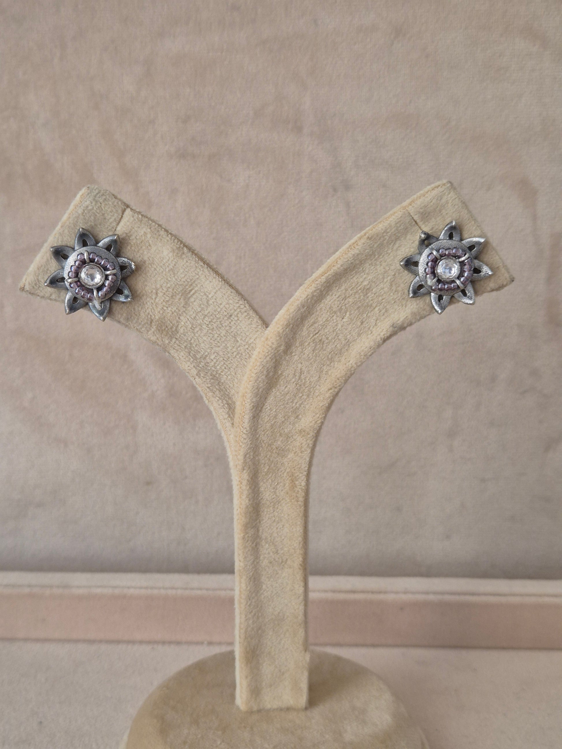 Pair of Neerja silver-toned floral stud earrings featuring an intricate starburst design with a central white stone on a stand. Naitri premium Neerja oxidized silver stud earrings from the Silver Jewellery collection by naitri.in