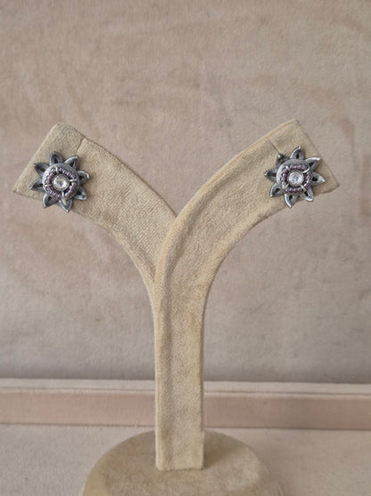 Pair of Neerja silver-toned floral stud earrings featuring an intricate starburst design with a central white stone on a stand. Naitri premium Neerja oxidized silver stud earrings from the Silver Jewellery collection by naitri.in