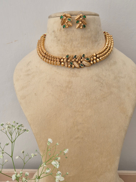 A gold-plated multi-strand choker necklace featuring a central green gemstone leaf motif, displayed on a beige mannequin bust with matching leaf stud earrings by naitri.in