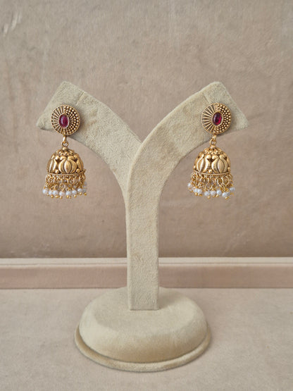 Pair of gold-plated Nivedita Jhumka earrings hanging on a beige velvet display stand, featuring textured circular studs with an oval pink gemstone center and bell-shaped drops fringed with small white pearls by naitri.in