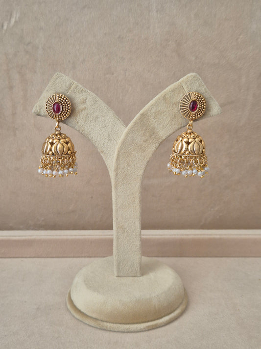 Pair of gold-plated Nivedita Jhumka earrings hanging on a beige velvet display stand, featuring textured circular studs with an oval pink gemstone center and bell-shaped drops fringed with small white pearls by naitri.in
