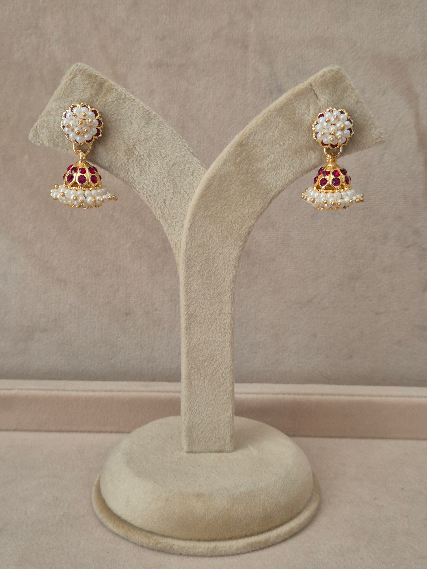 Pair of gold-plated Niyati Jhumka earrings hanging on a beige velvet display stand, featuring circular studs packed with tiny white seed pearls and traditional bell-shaped drops adorned with a band of red gemstones and a fringe of dangling pearls by naitri.in