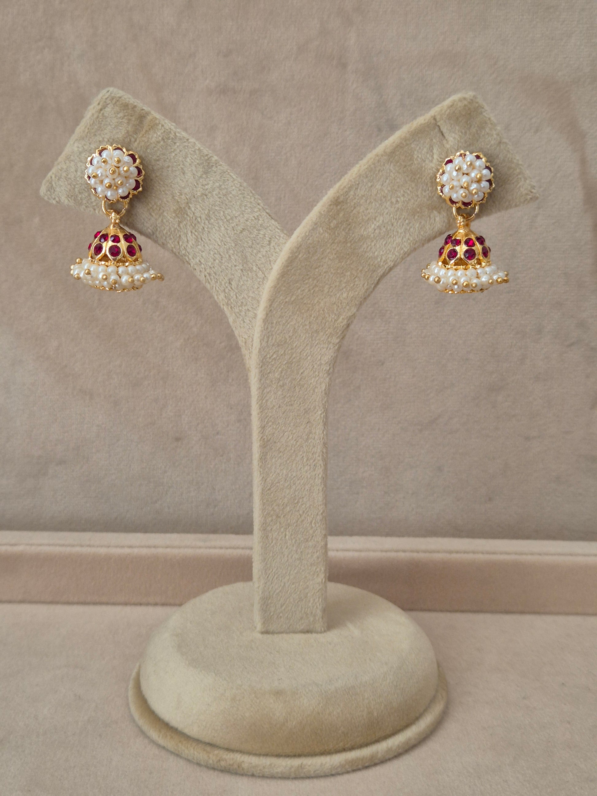 Pair of gold-plated Niyati Jhumka earrings hanging on a beige velvet display stand, featuring circular studs packed with tiny white seed pearls and traditional bell-shaped drops adorned with a band of red gemstones and a fringe of dangling pearls by naitri.in