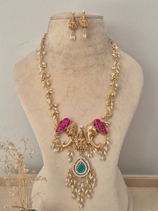 Long gold temple necklace featuring magenta stone-encrusted elephant motifs, a green teardrop center pendant with dangling pearls, with matching earrings by naitri.in
