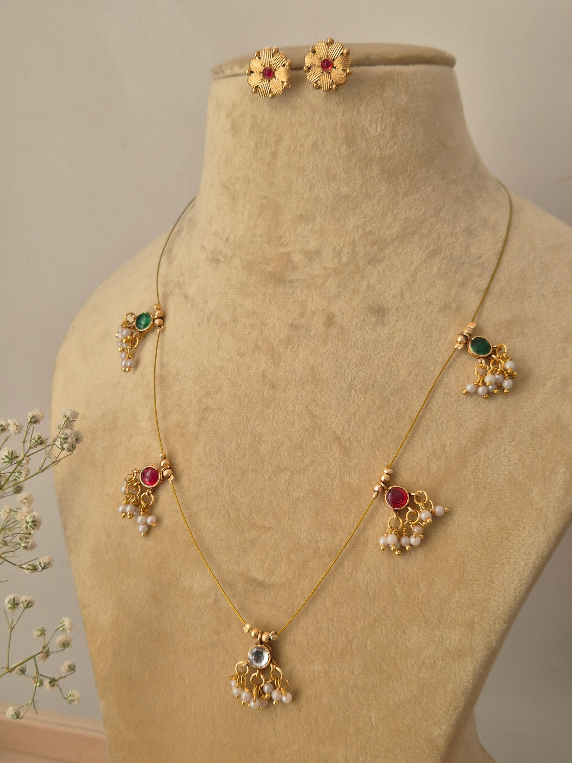 Minimalist gold necklace with five hanging stone pendants in emerald green and ruby red, paired with small round floral-patterned stud earrings.