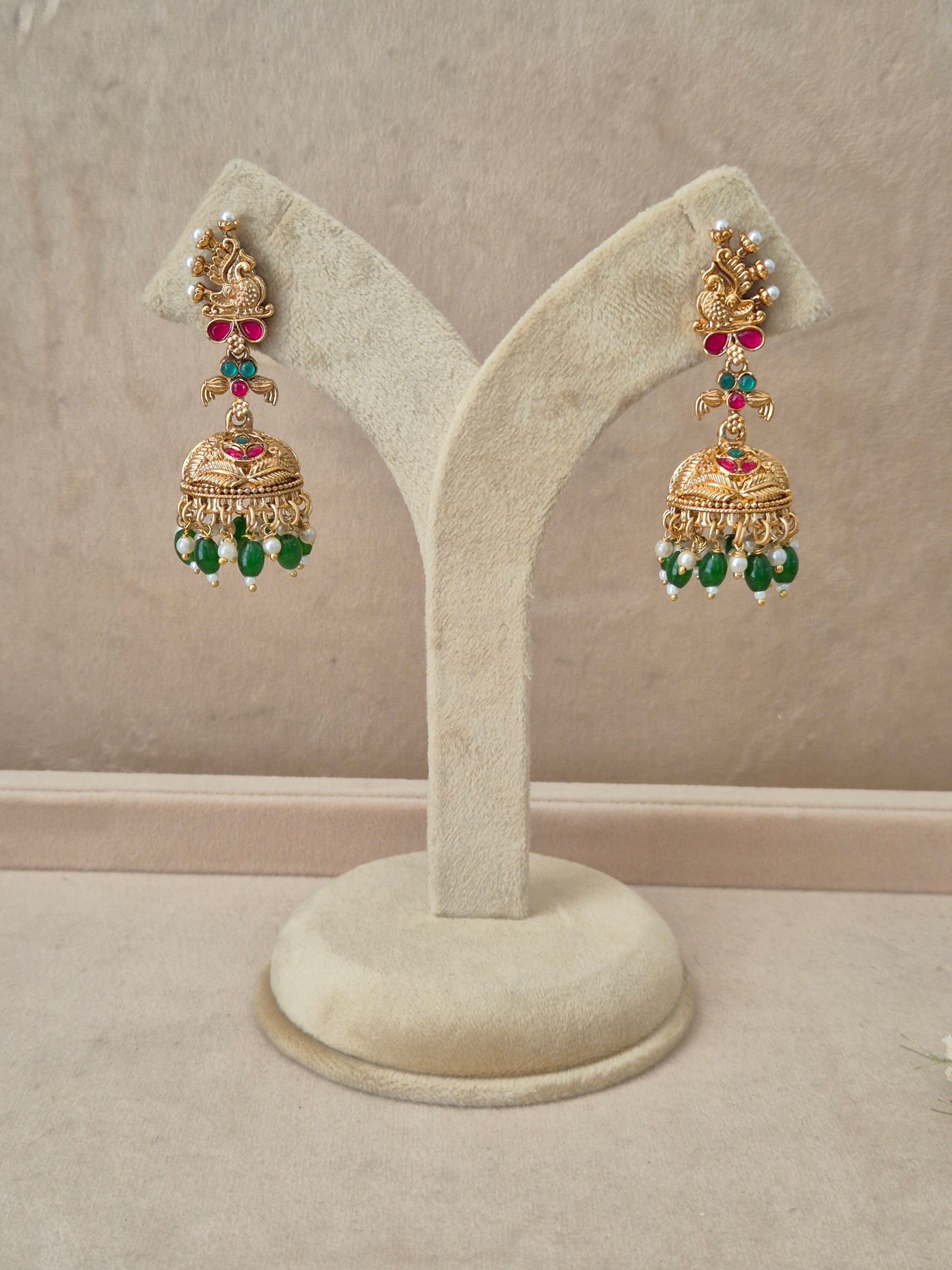 Pair of gold-plated Oindrila Jhumka earrings hanging on a beige velvet display stand, featuring detailed peacock motif studs with red and green gemstone accents and traditional bell-shaped drops fringed with small green beads by naitri.in