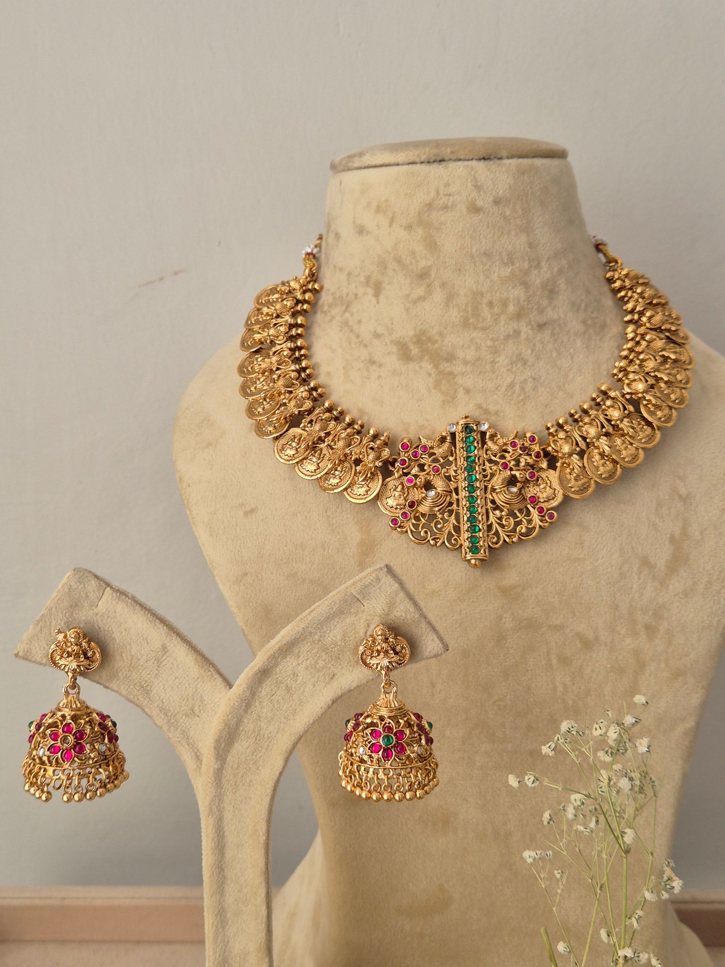 Gold-toned traditional Indian temple jewelry set featuring a choker-style necklace with coin motifs and a central rectangular pendant detailed with red and green gemstone accents. Paired with matching hanging jhumka earrings featuring pink/red bead embellishments, by naitri.in