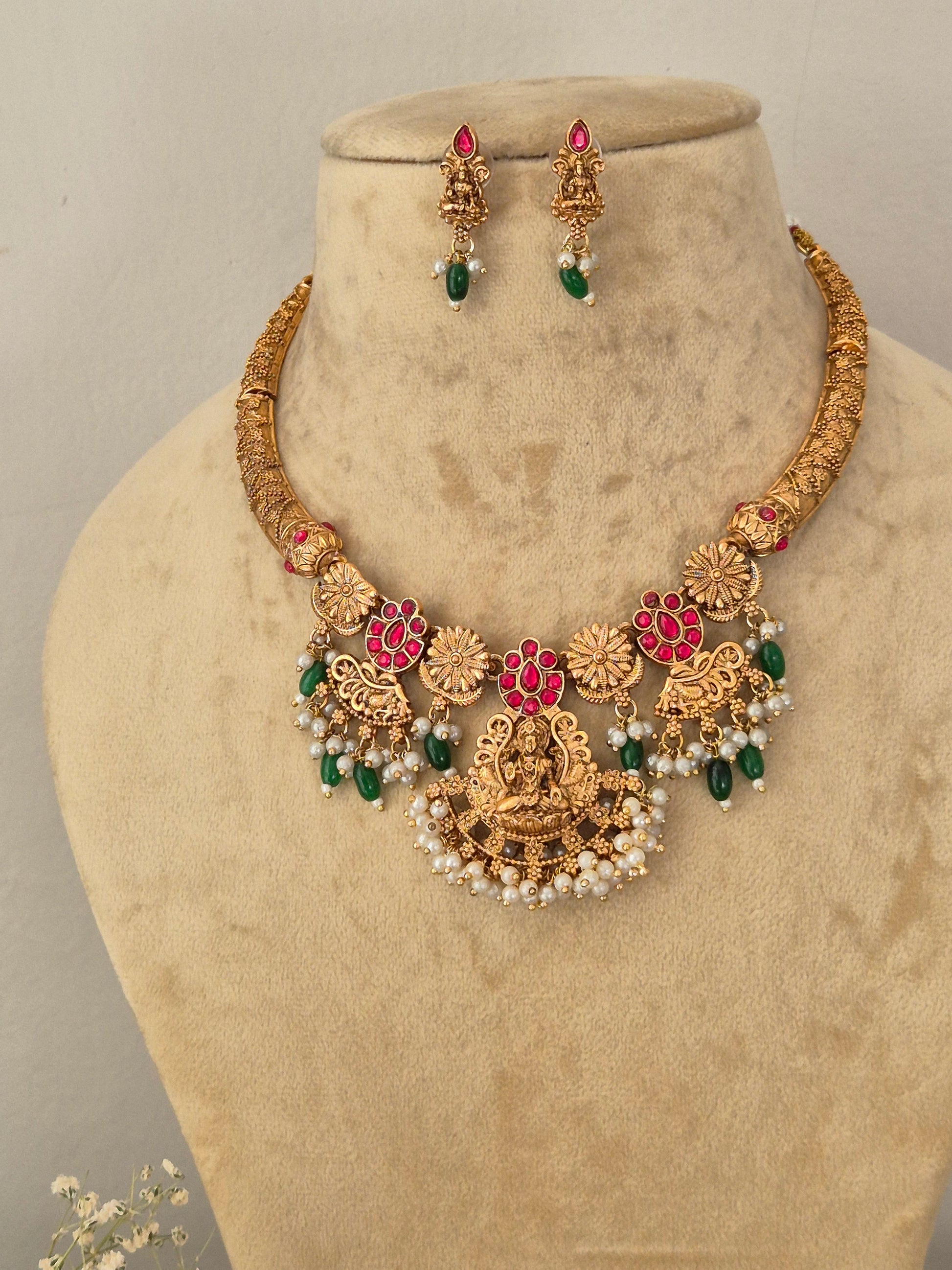 Gold-toned traditional Indian jewelry set featuring a necklace and matching earrings the necklace has intricate floral motifs with red enamel, green stone accents, and white pearl-like beads by naitri.in