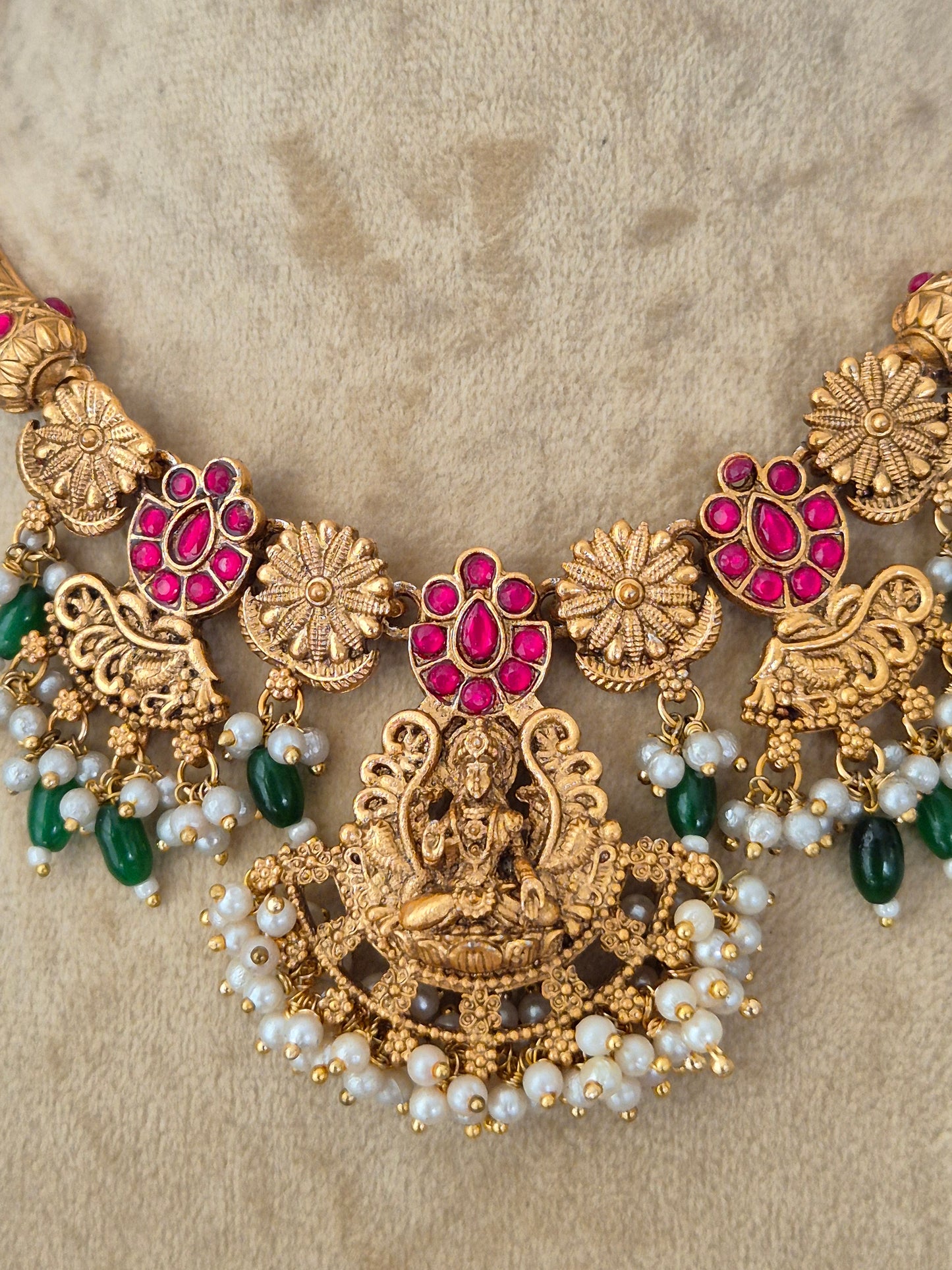An ornate gold-toned traditional necklace with red gemstone floral motifs, white pearl bead strands, and green bead accents, necklace is designed with intricate floral metalwork, red enamel accents, green gemstones, and a fringe of hanging white pearl-like beads by naitri.in