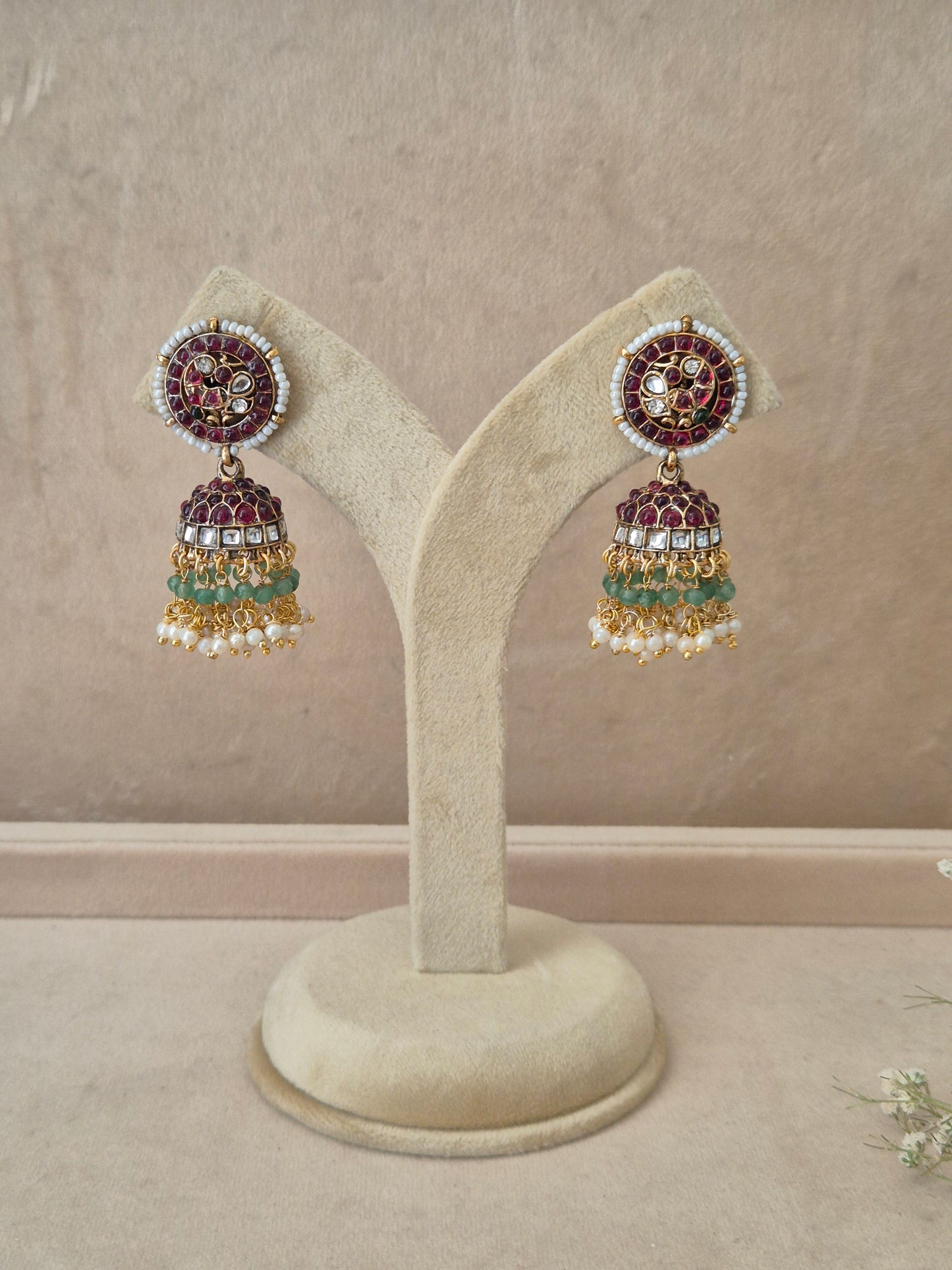 Pair of gold-plated Poorvi Jhumka earrings hanging on a beige velvet display stand, featuring round floral studs inlaid with dark pink enamel and bell-shaped drops adorned with rows of red stones, green beads, and white pearls by naitri.in