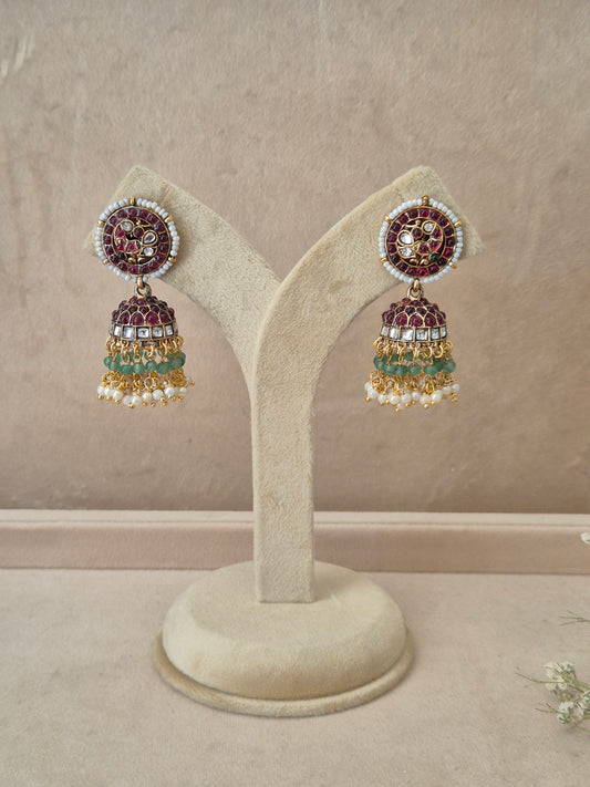Pair of gold-plated Poorvi Jhumka earrings hanging on a beige velvet display stand, featuring round floral studs inlaid with dark pink enamel and bell-shaped drops adorned with rows of red stones, green beads, and white pearls by naitri.in