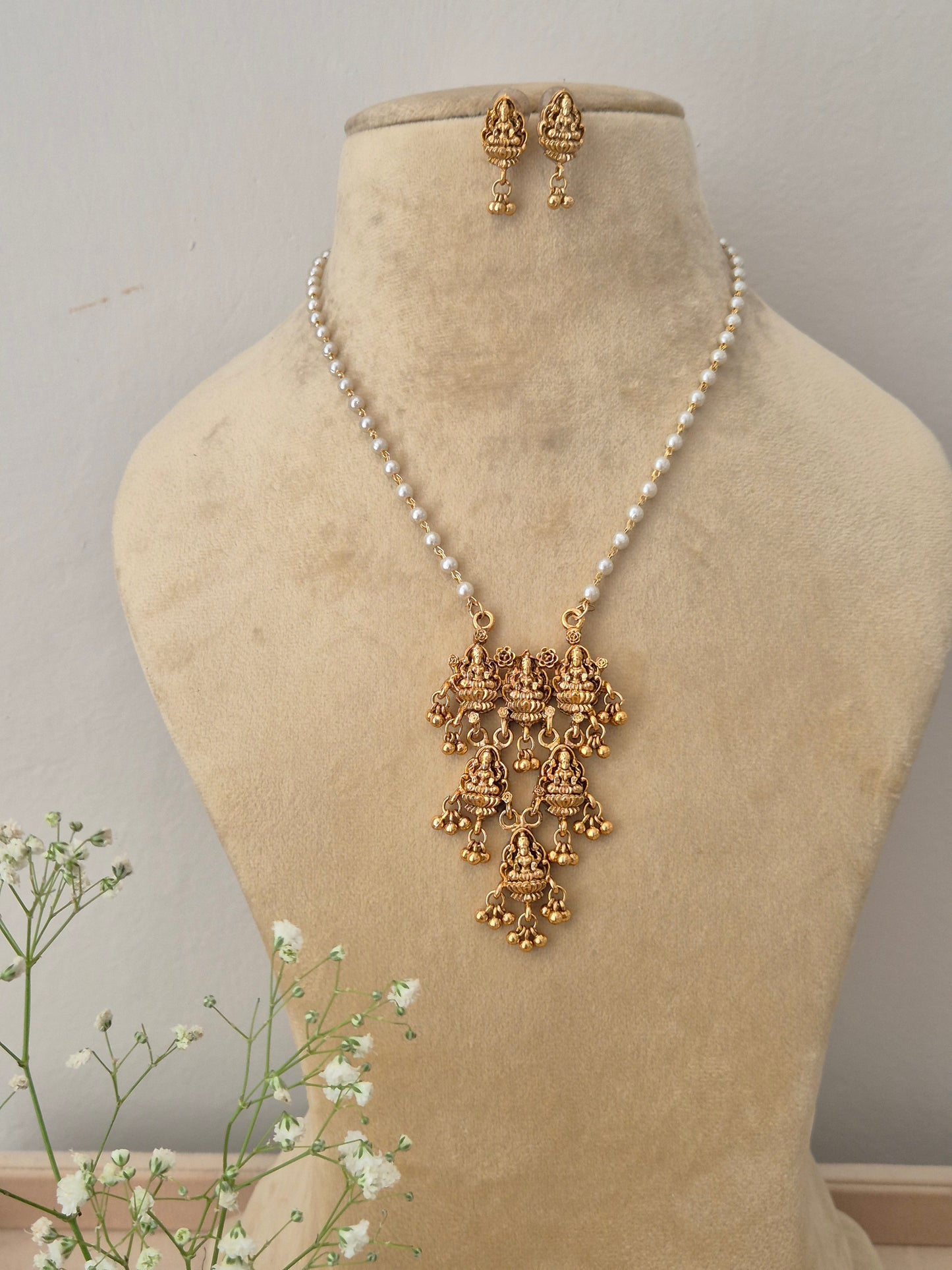 A gold-plated multi-tiered temple jewelry pendant featuring intricate traditional goddess engravings, suspended on a single strand pearl chain with matching earrings by naitri.in