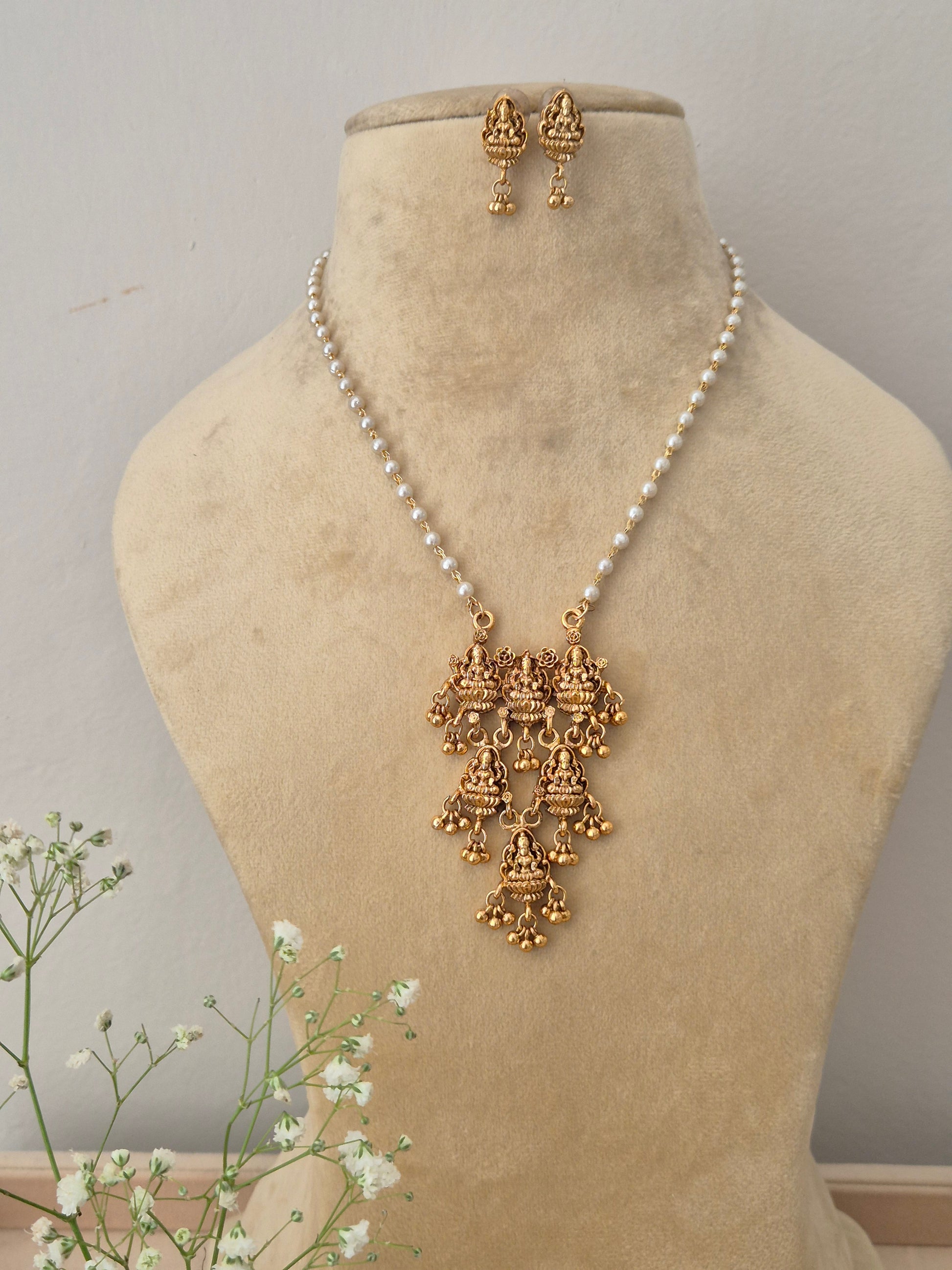 A gold-plated multi-tiered temple jewelry pendant featuring intricate traditional goddess engravings, suspended on a single strand pearl chain with matching earrings by naitri.in