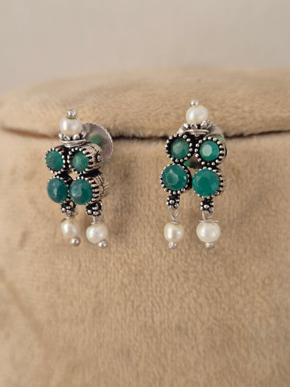 A pair of traditional Indian jhumka earrings with silver-toned metalwork, deep green round stones, and white pearl beads, by naitri.in