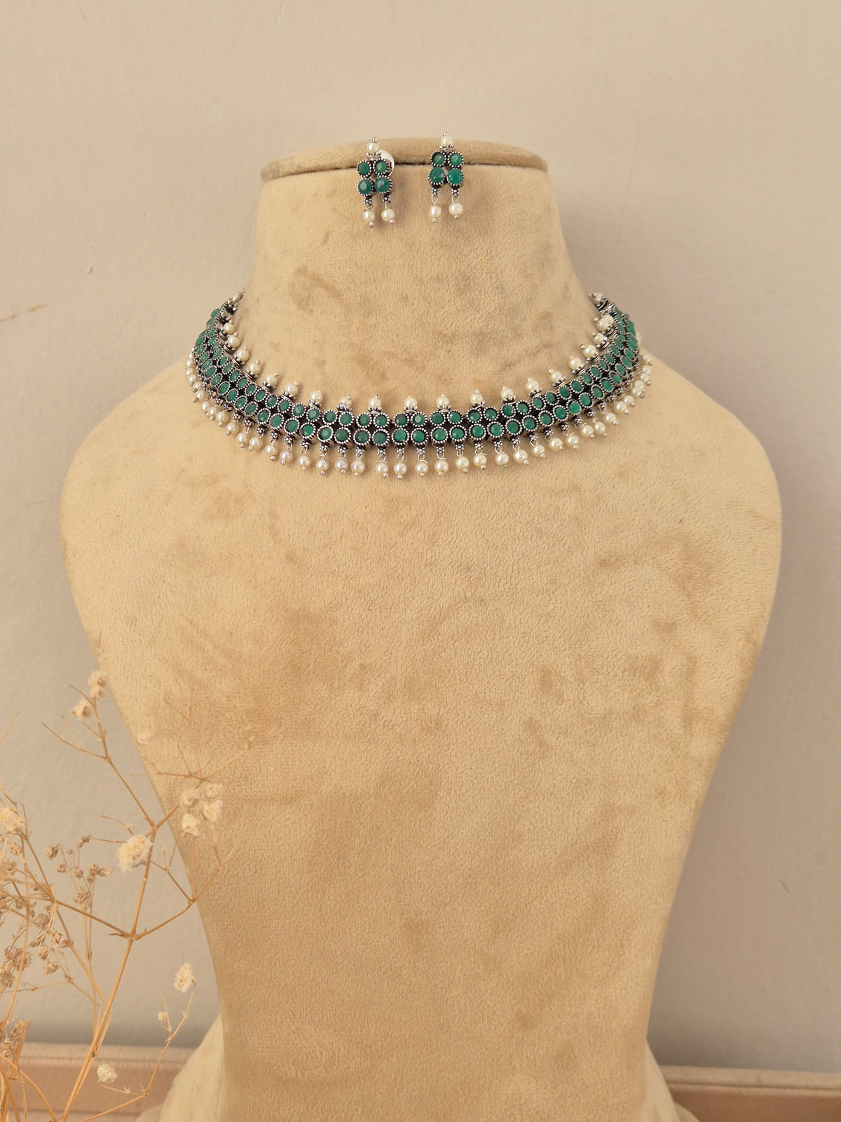 A traditional Indian jewelry set featuring a choker necklace and matching earrings the jewelry features silver-toned metal work, deep green stones, and white faux pearls by naitri.in