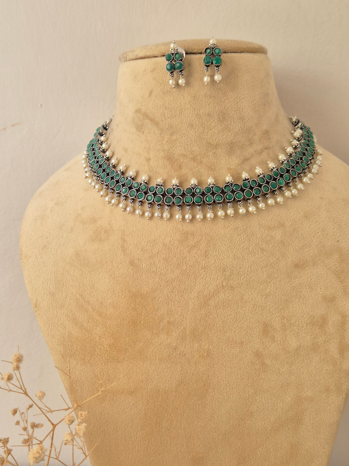 A traditional Indian jewelry set featuring a choker necklace and matching earrings. The jewelry features silver-toned metalwork, deep green stones, and white pearl bead accents by naitri.in
