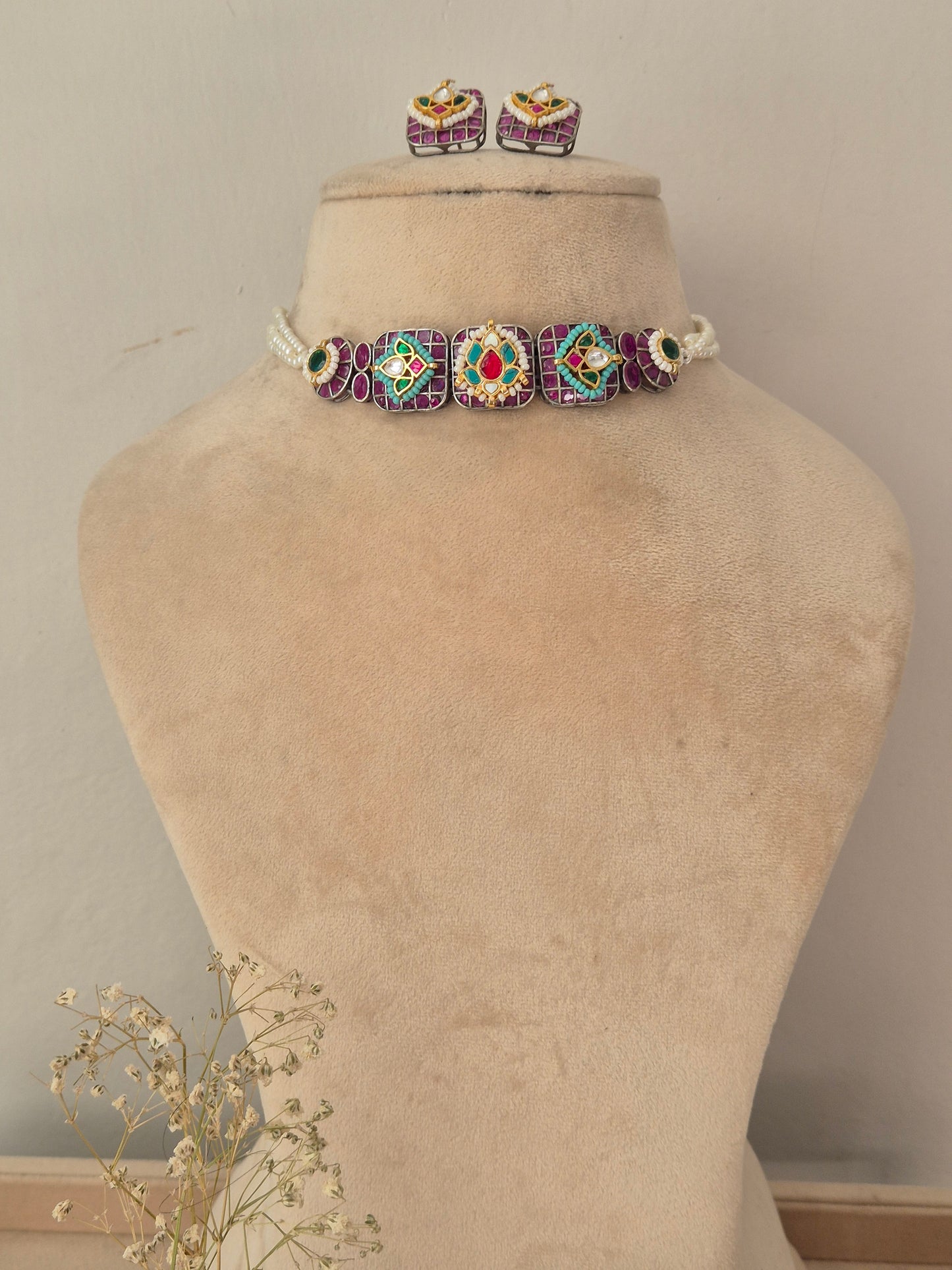 A jewelry set featuring a beaded choker necklace with matching stud earrings. The jewelry has a white base with purple, green, and red beaded geometric patterns. by naitri.in