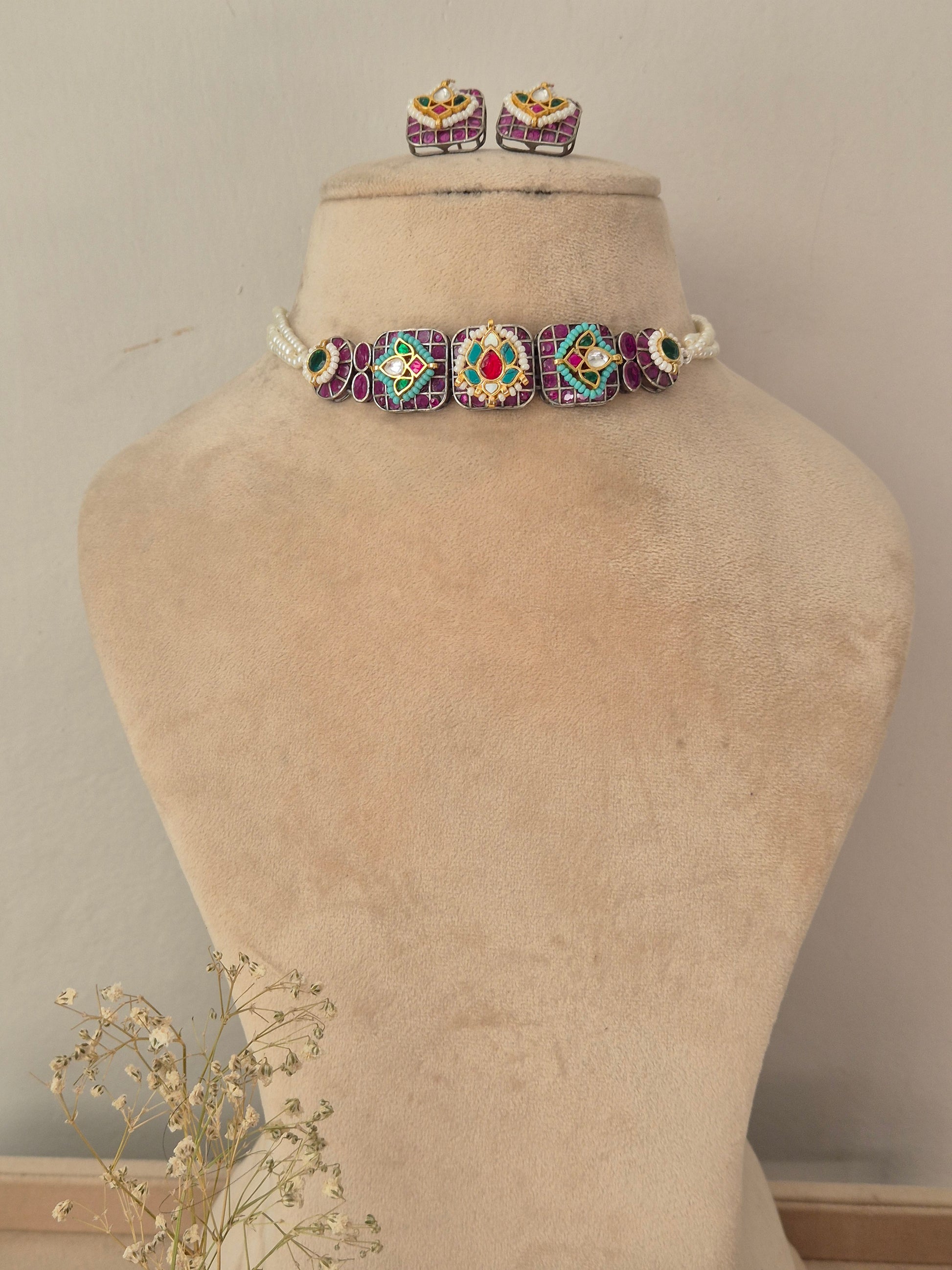 A jewelry set featuring a beaded choker necklace with matching stud earrings. The jewelry has a white base with purple, green, and red beaded geometric patterns. by naitri.in