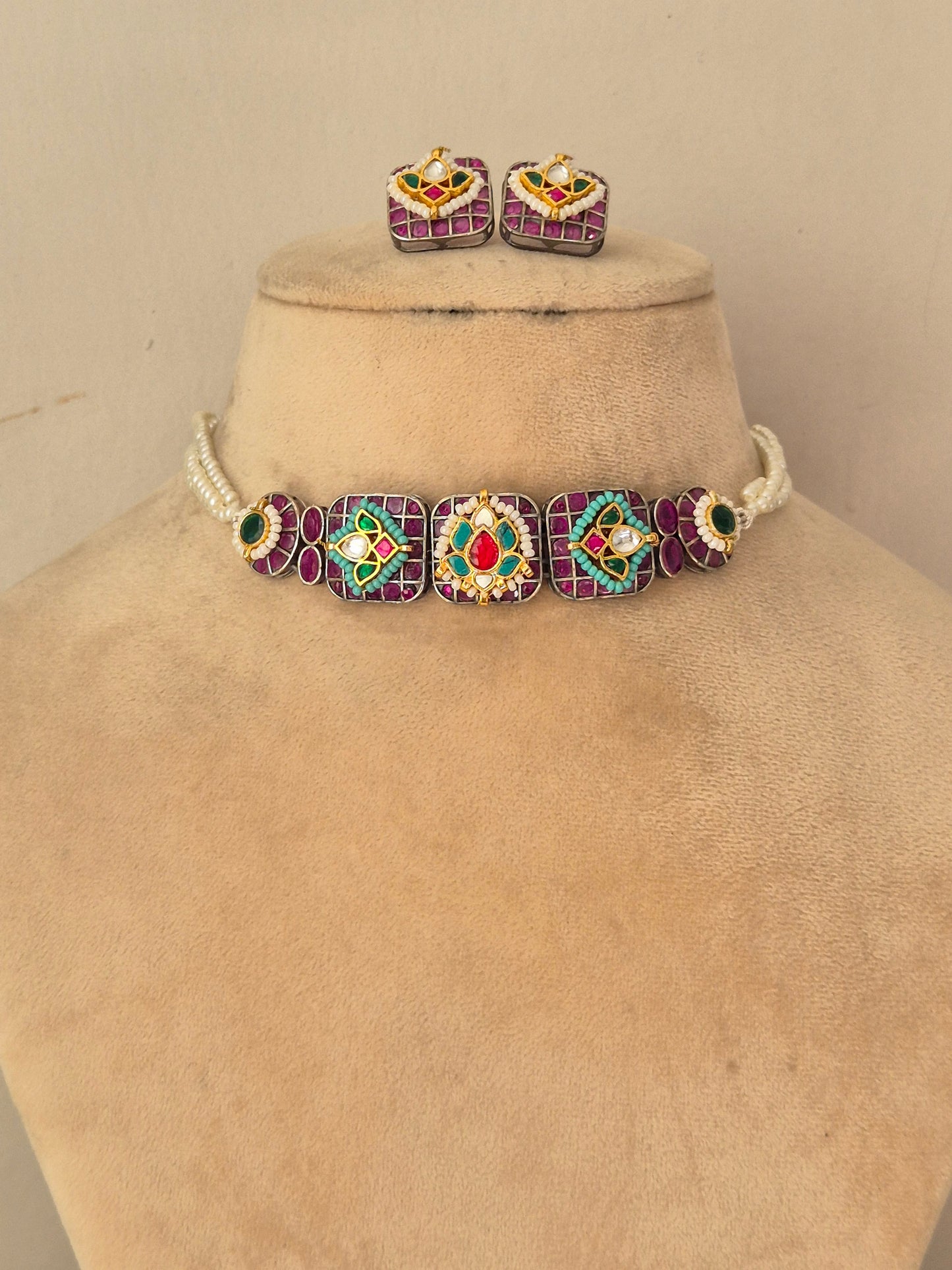 A jewelry set, featuring a choker necklace and matching stud earrings. The necklace has a white beaded base with four large square panels in purple, green, red, and gold, while the matching earrings have a similar multicolor design with a central gold heart by naitri.in