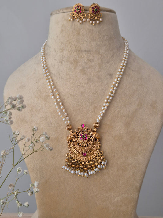 A gold-plated temple jewelry pendant featuring a goddess motif and pink stone accents, suspended from a double-strand white pearl chain with matching stud earring by naitri.in