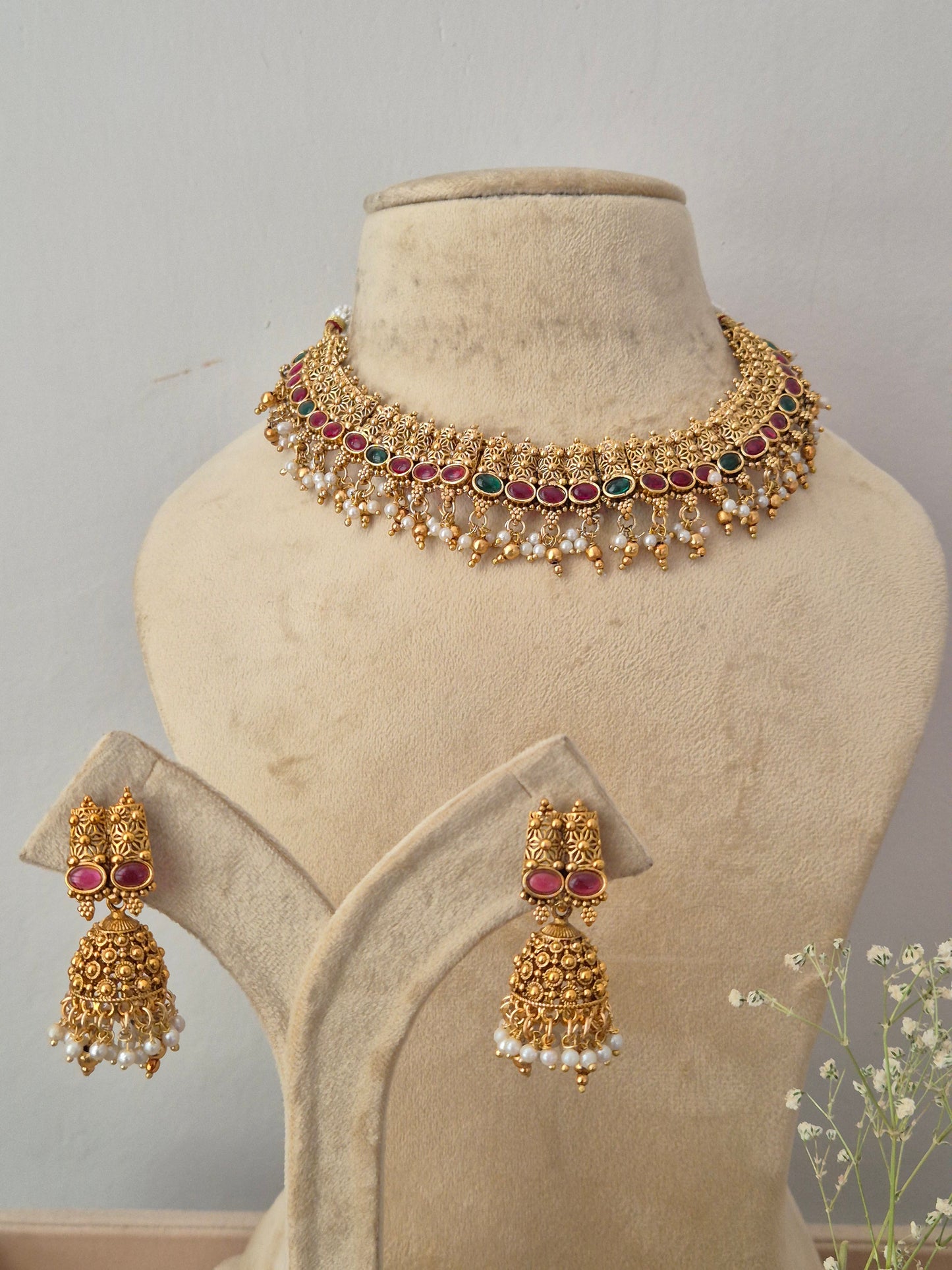 Gold-plated Indian choker necklace and matching dangle earrings,  the necklace features intricate traditional motifs, a row of red and green gemstones, and a fringe of hanging white faux pearls, with matching pink stone and pearl drop earrings by naitri.in