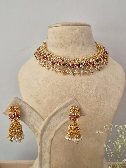 Gold-plated Indian choker necklace and matching dangle earrings,  the necklace features intricate traditional motifs, a row of red and green gemstones, and a fringe of hanging white faux pearls, with matching pink stone and pearl drop earrings by naitri.in