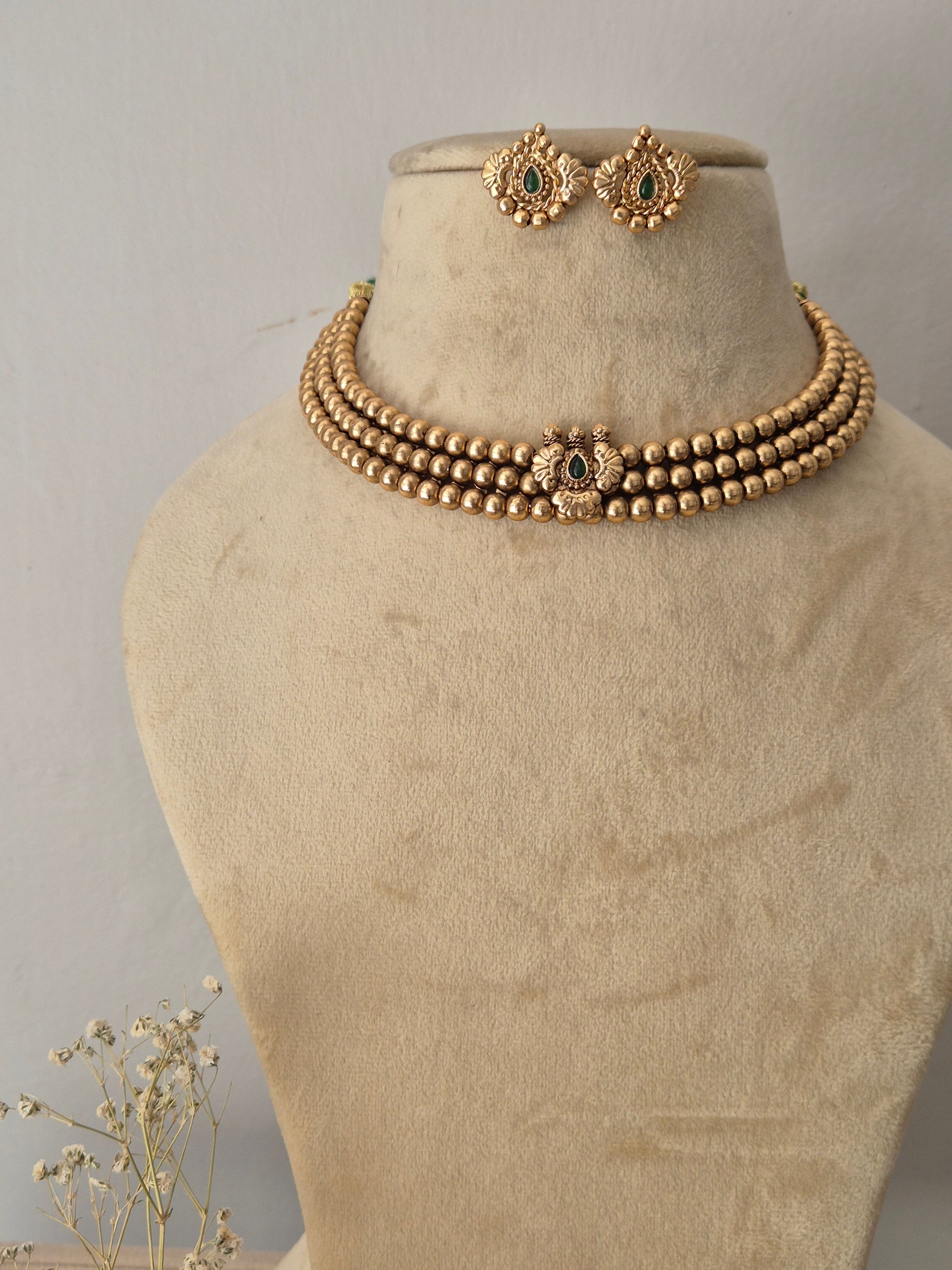 Gold-toned traditional Indian necklace set featuring a multi-layered beaded collar choker with a central peacock motif and green gemstone accent, paired with matching round stud earrings by naitri.in