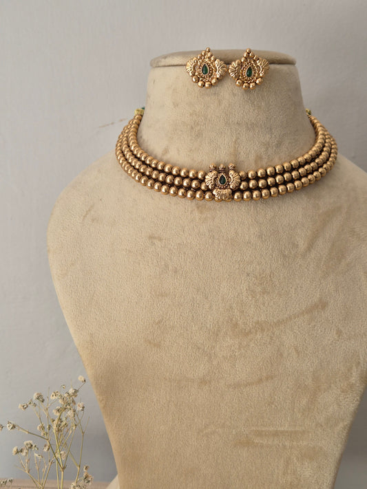 Gold-toned traditional Indian necklace set featuring a multi-layered beaded collar choker with a central peacock motif and green gemstone accent, paired with matching round stud earrings by naitri.in