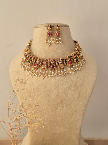 Raina Gold Temple Choker Set displayed on a beige mannequin, showcasing intricate traditional craftsmanship paired with multi-colored gemstone embellishments and dangling pearl drops by Naitri.in