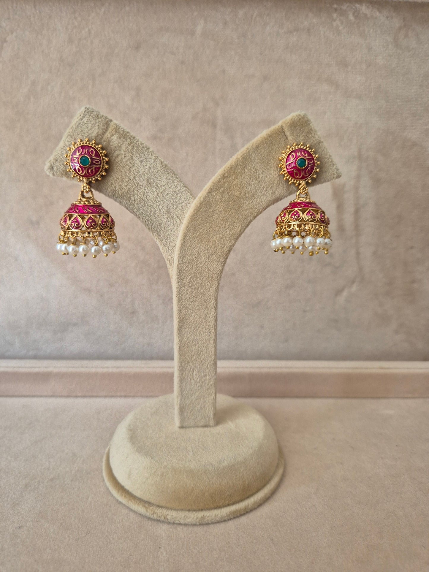Pair of gold-plated Rakshita Jhumka earrings hanging from a beige velvet display stand, featuring round floral studs detailed with dark pink and green enamel work and traditional bell-shaped drops fringed with small white seed pearls. by naitri.in
