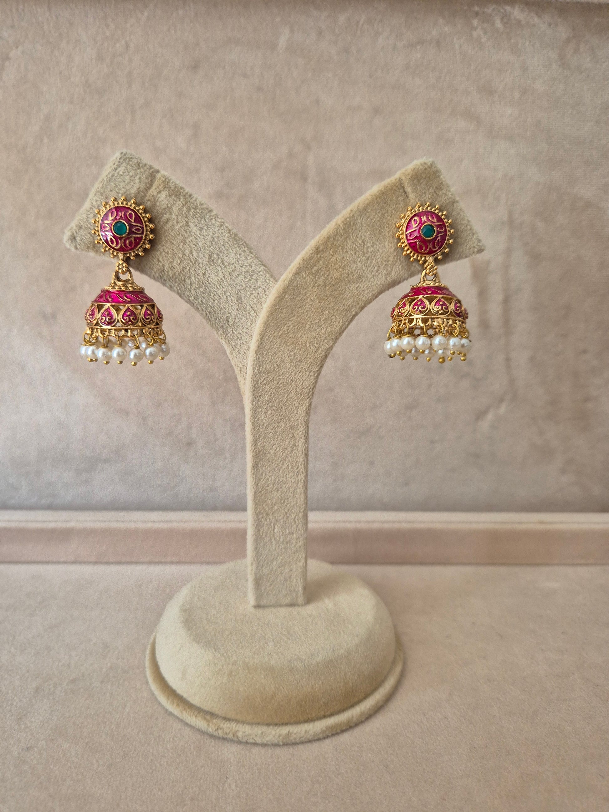 Pair of gold-plated Rakshita Jhumka earrings hanging from a beige velvet display stand, featuring round floral studs detailed with dark pink and green enamel work and traditional bell-shaped drops fringed with small white seed pearls. by naitri.in