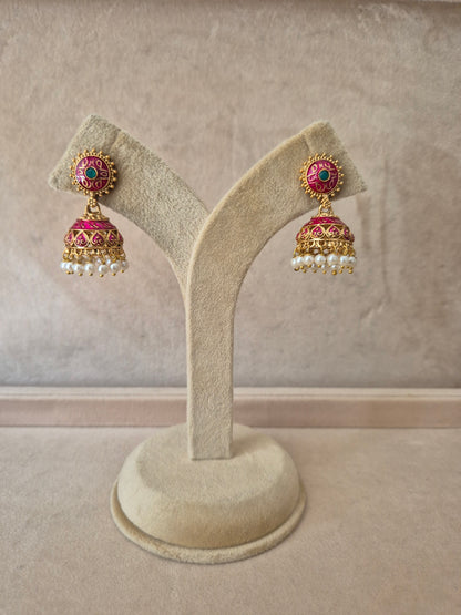 Pair of gold-plated Rakshita Jhumka earrings hanging from a beige velvet display stand, featuring round floral studs detailed with dark pink and green enamel work and traditional bell-shaped drops fringed with small white seed pearls. by naitri.in