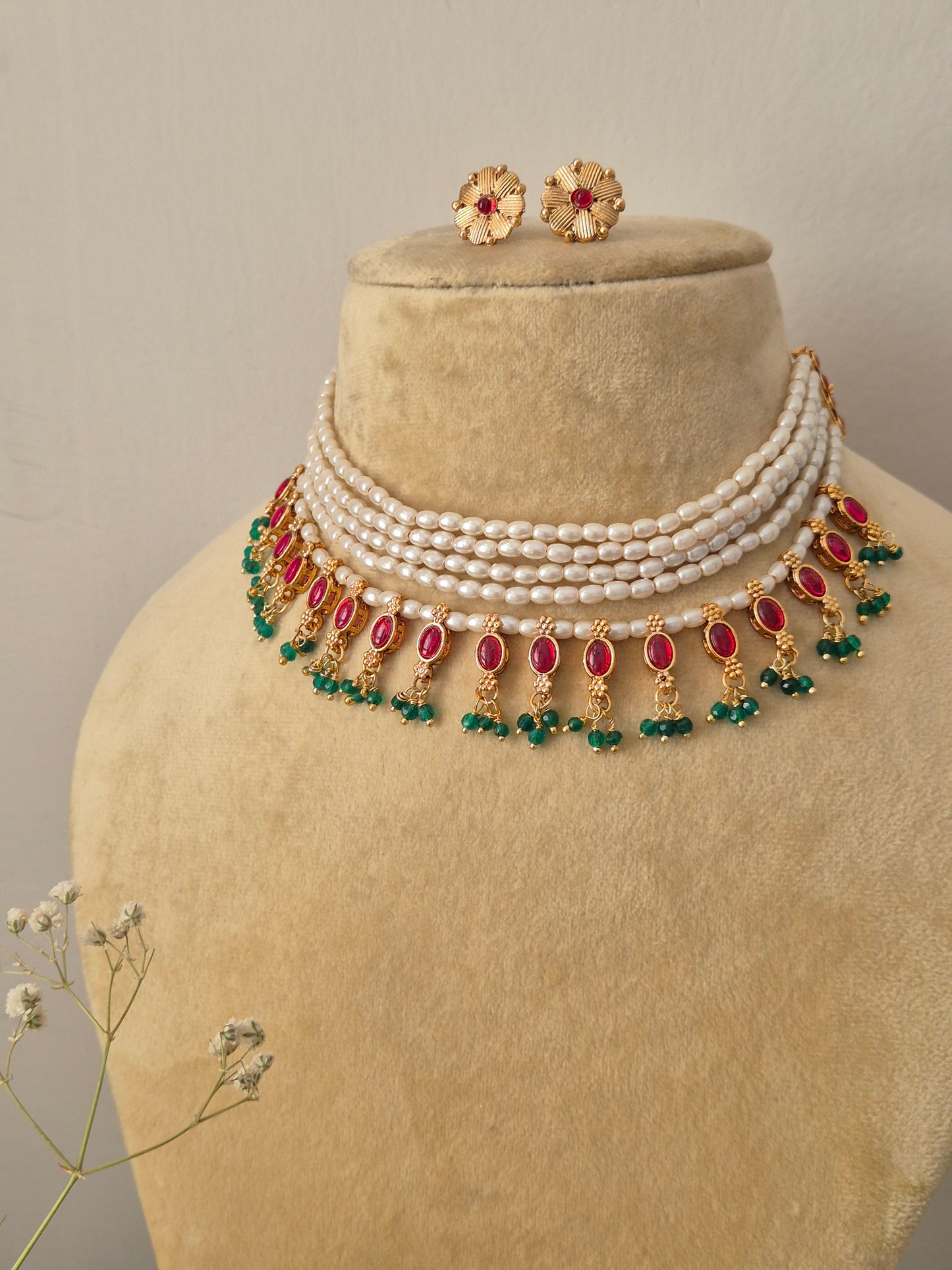 Gold-polished layered pearl choker with red and green stone embellishments and coordinating textured stud earrings Handcrafted multi-strand pearl choker necklace and matching gold earrings featuring red stone drops and green bead fringes by naitri.in