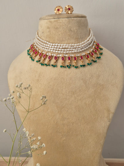 Handcrafted choker necklace featuring a four-layer white pearl strand collar, dangling teardrop ruby-red stones, and emerald-green bead cluster fringes with matching round gold stud earrings by naitri.in
