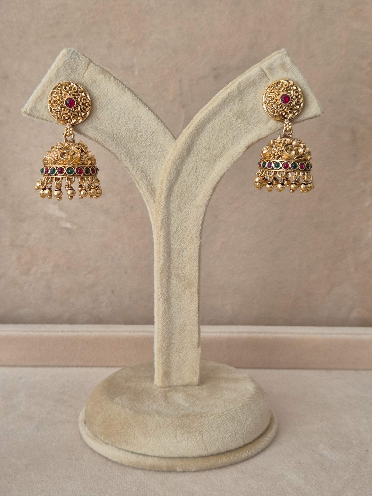 Pair of traditional gold Jhumka earrings with intricate detailing and pink gemstones, displayed on a white jewelry stand by naitri.in