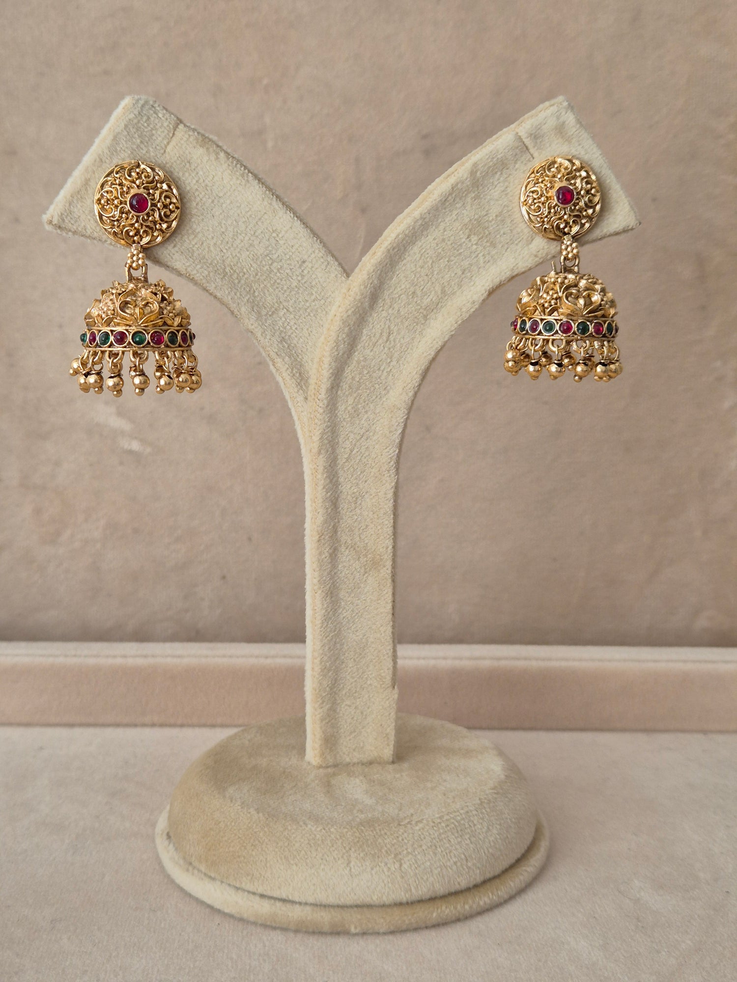 Pair of traditional gold Jhumka earrings with intricate detailing and pink gemstones, displayed on a white jewelry stand by naitri.in
