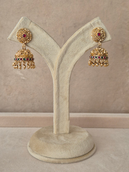 Pair of traditional gold Jhumka earrings with intricate detailing and pink gemstones, displayed on a white jewelry stand by naitri.in