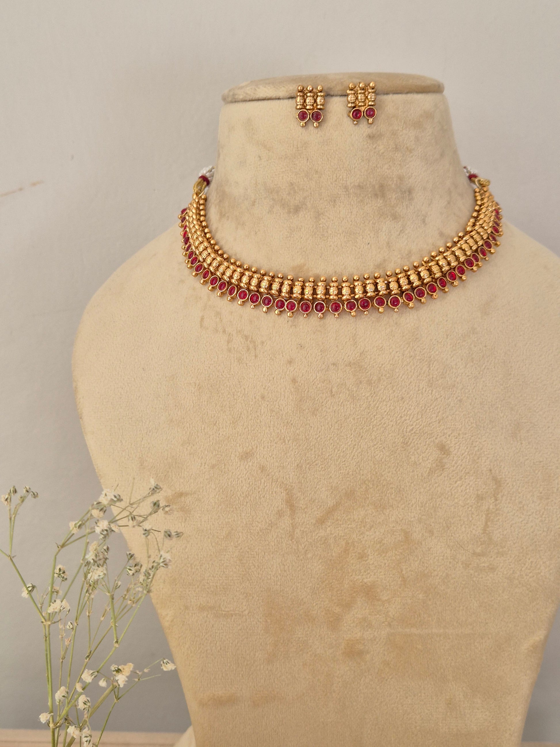 Gold-toned traditional Indian choker necklace and matching small drop earrings set, it is designed with a row of small vertical bars leading down to a dense row of deep pink or ruby red gemstones, by naitri.in