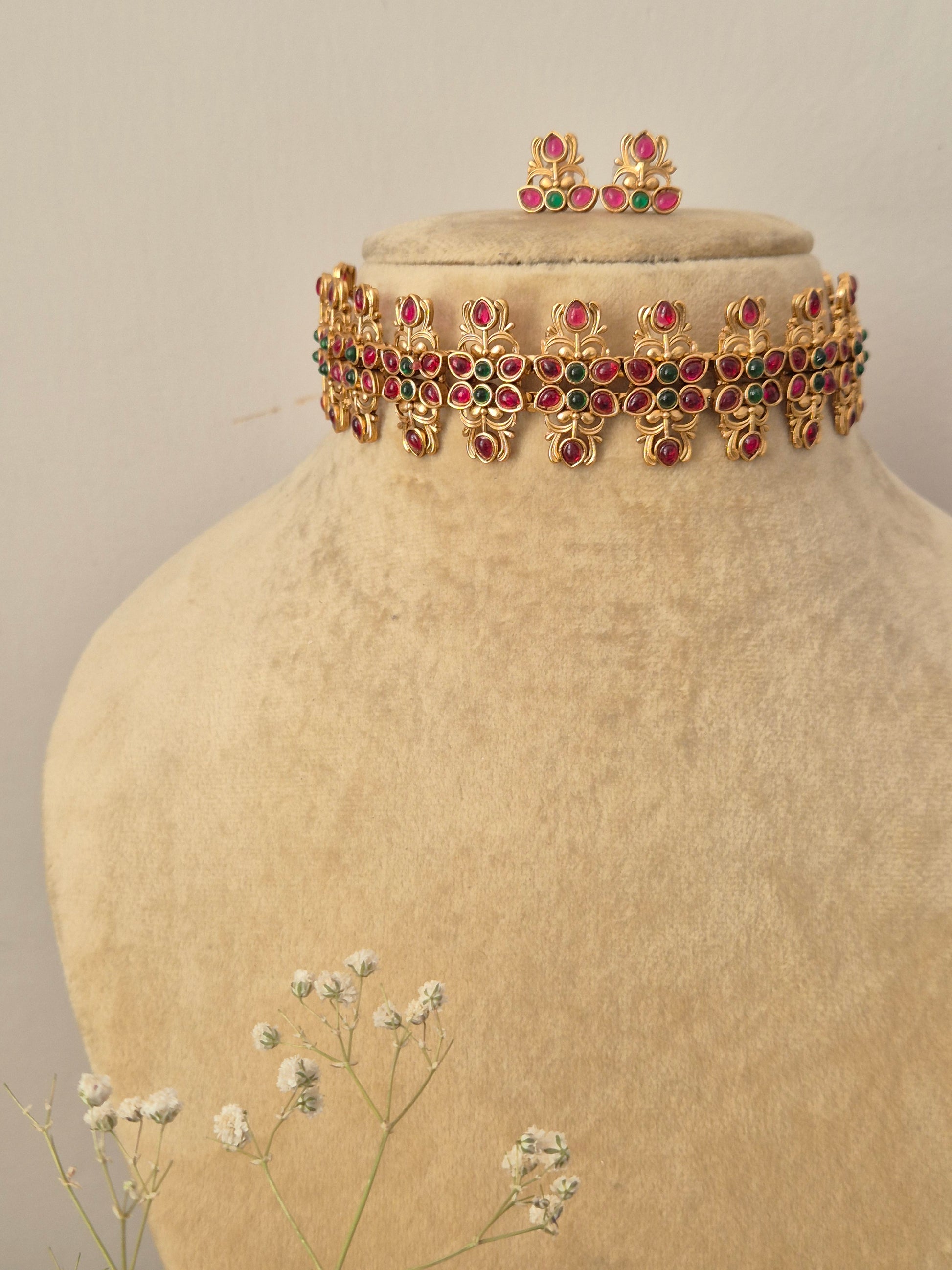 Handcrafted gold-plated temple jewelry choker necklace featuring a repeating pattern of ruby-red and emerald-green kemp stones in floral motifs, paired with matching small stud earrings by naitri.in