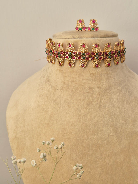Handcrafted gold-plated temple jewelry choker necklace featuring a repeating pattern of ruby-red and emerald-green kemp stones in floral motifs, paired with matching small stud earrings by naitri.in