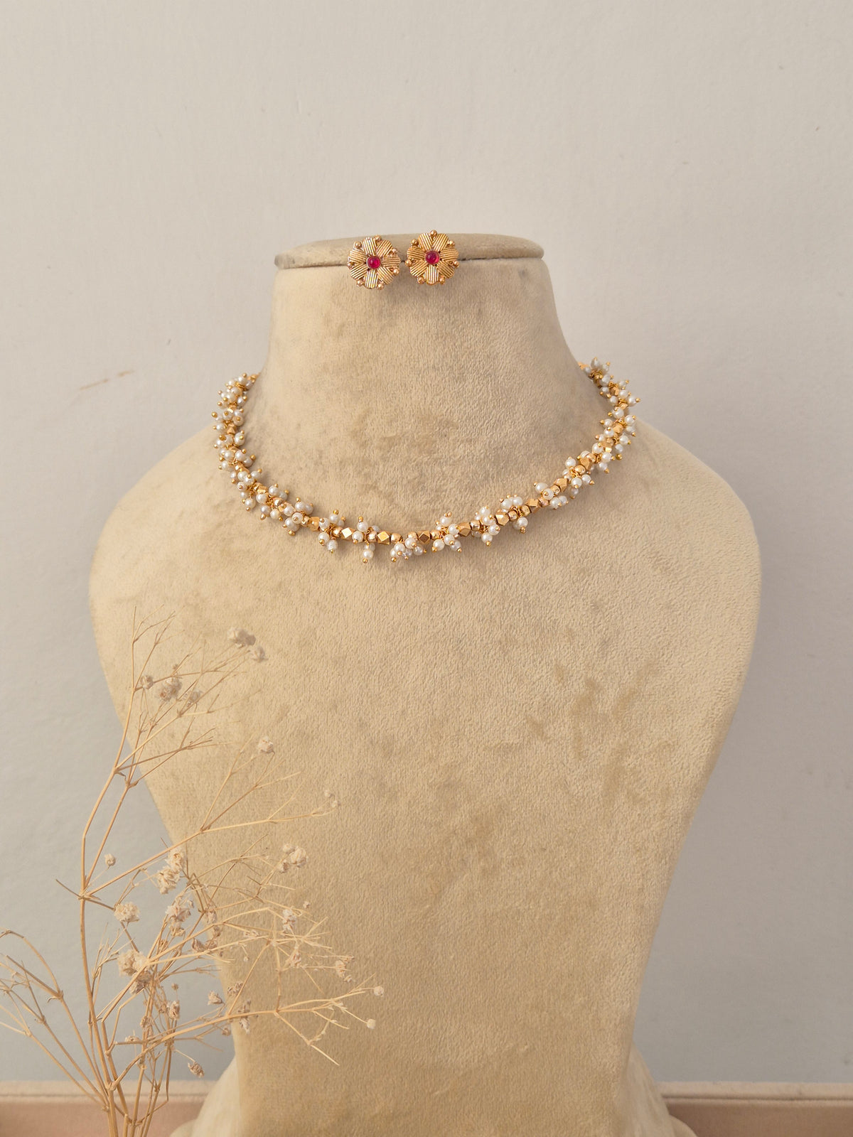 Ruby Necklace Set displayed on a neutral beige mannequin, featuring a delicate golden chain intertwined with vibrant red ruby-like stone accents and matching stud earrings by Naitri.in