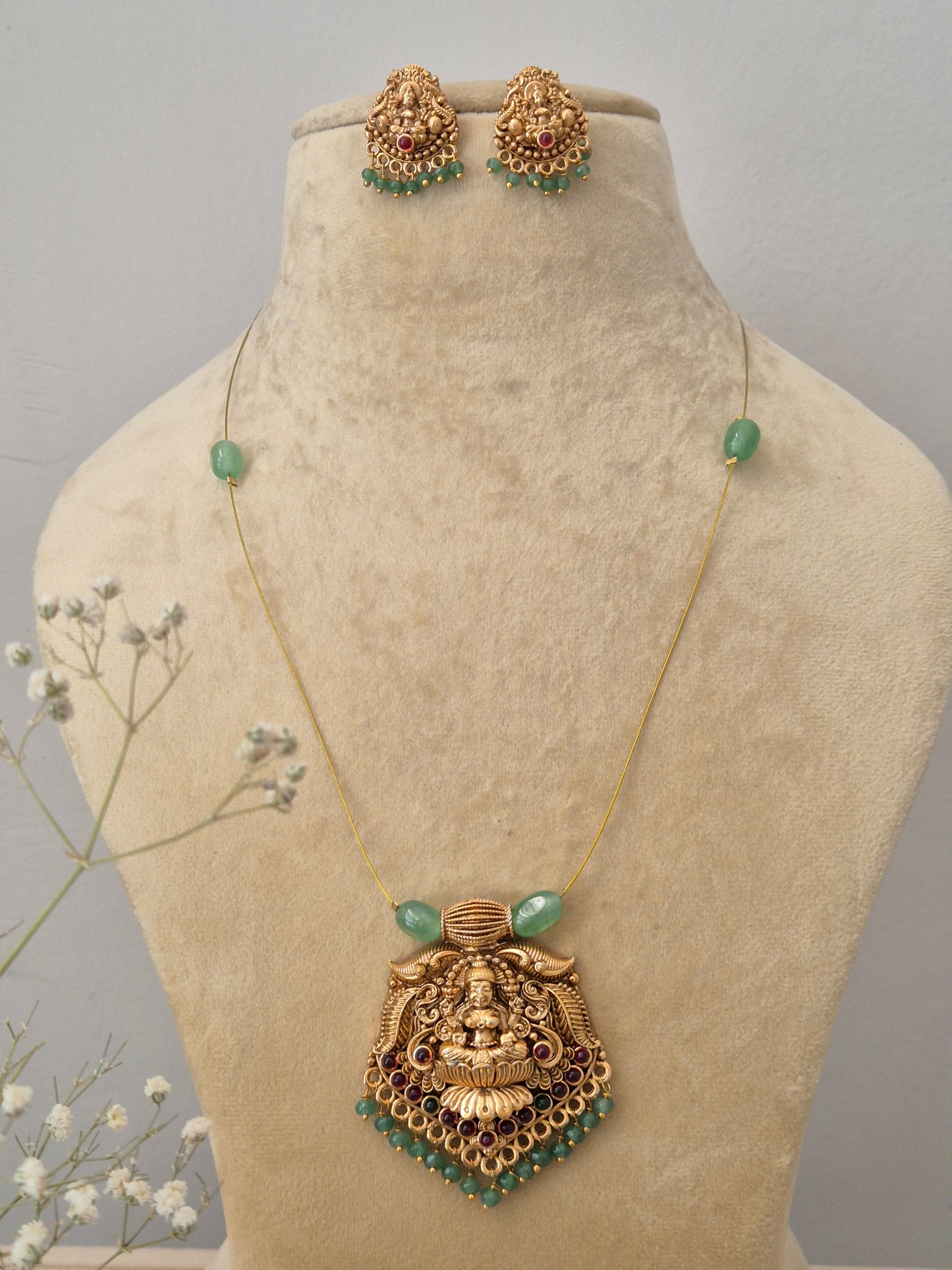 Antique gold-finish Temple jewelry set featuring a pendant with an intricate deity carving, green bead accents, an invisible chain, and matching jhumka-style earrings by naitri.in