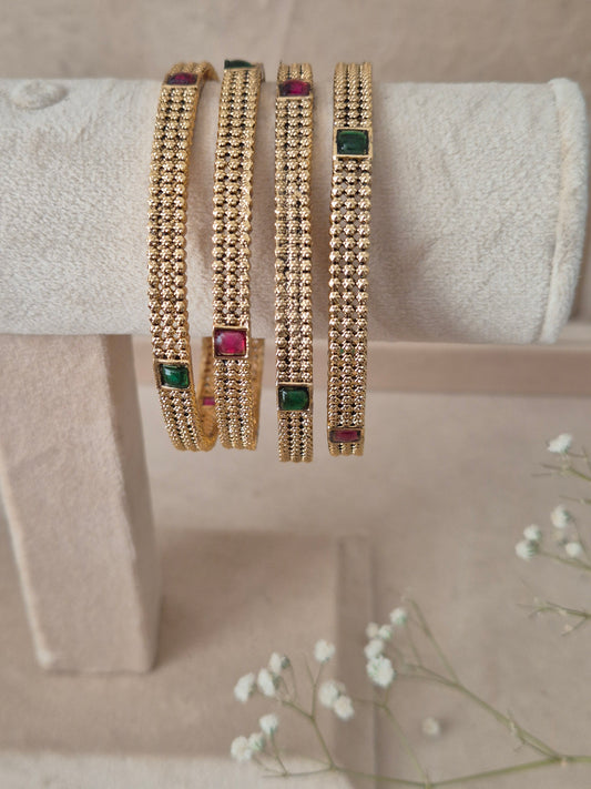 Set of 4 Savya gold bangles by Naitri featuring intricate textured metal engravings, accented with alternating ruby-red and emerald-green gemstones, by naitri.in