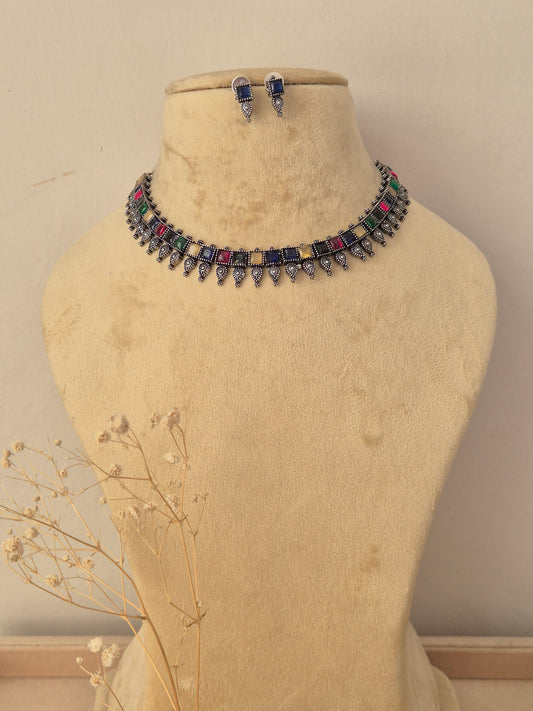 A silver-toned choker necklace and matching drop earrings  The necklace features a row of colorful rectangular gemstones in blue, pink, green, yellow, and red, with dangling silver teardrop-shaped charms by naitri.in