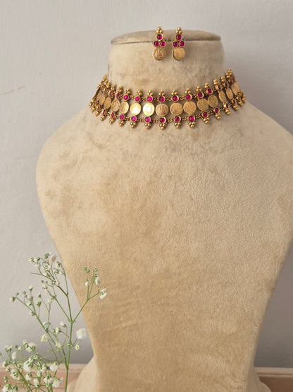 A gold-plated traditional ethnic choker necklace featuring a row of circular coin motifs and hanging red bead droplets, with matching earrings by naitri.in