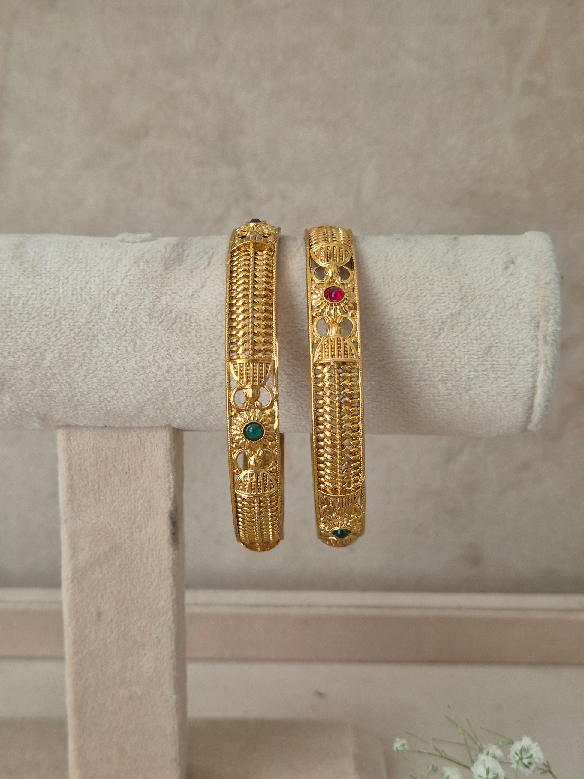 Two gold bangles with intricate filigree and small red and green stone accents, by naitri.in