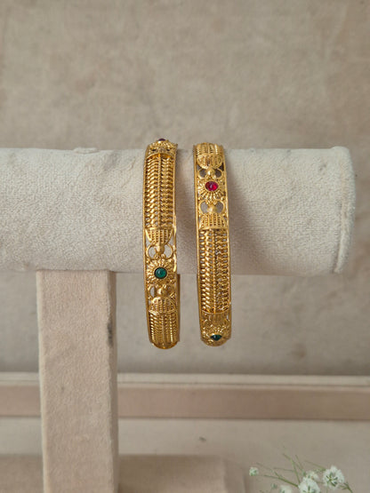 Two gold bangles with intricate filigree and small red and green stone accents, by naitri.in